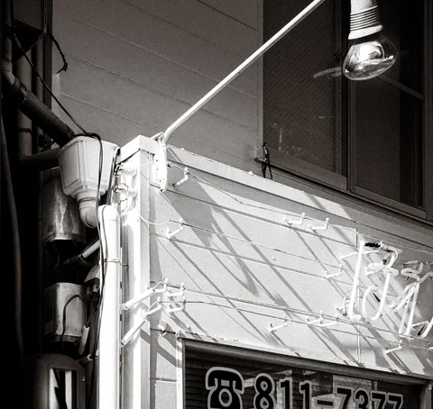 An overhead lamp and tangled sign framework cast angular shadows across a pale wall in black and white.