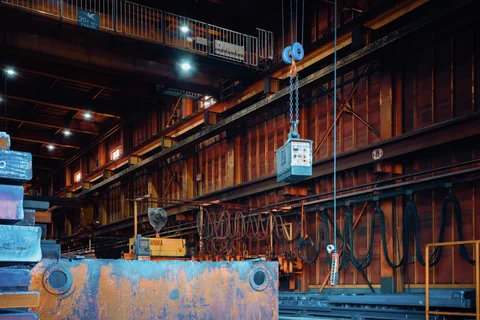 A blue hoist hook and chain hang inside a rust-colored industrial hall with illuminated beams and heavy equipment.
