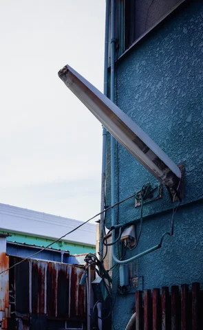 A long pale light fixture angles upward from a blue wall beside exposed wires and pipes.