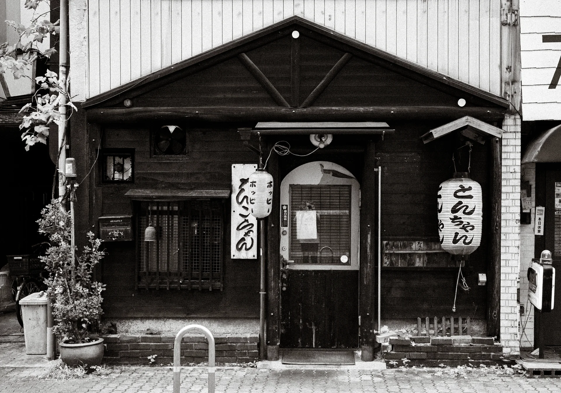 Two white paper lanterns flank the doorway of a dark wooden storefront in a black and white view.