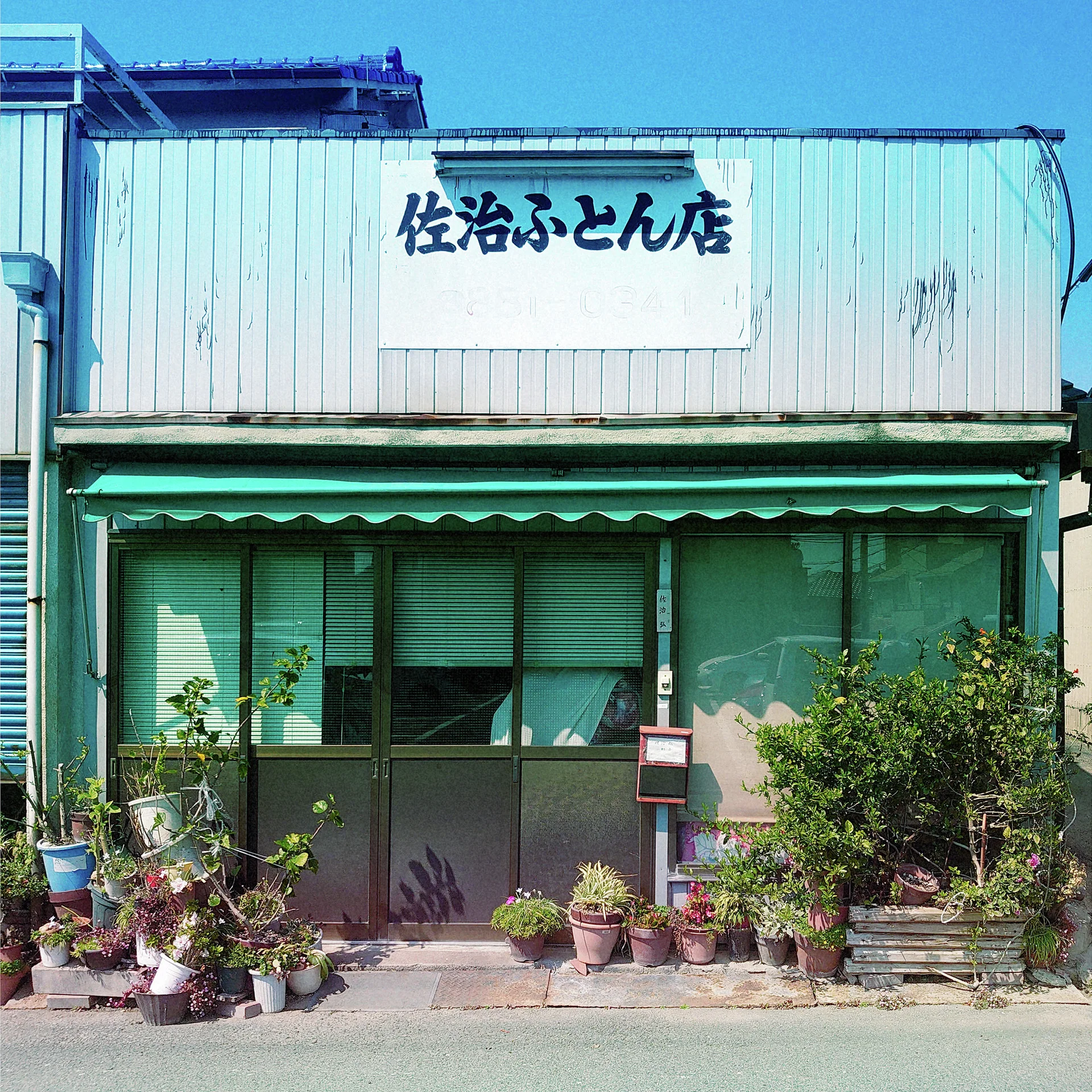 Potted plants line a low corrugated shop beneath a green awning and a white Japanese sign.