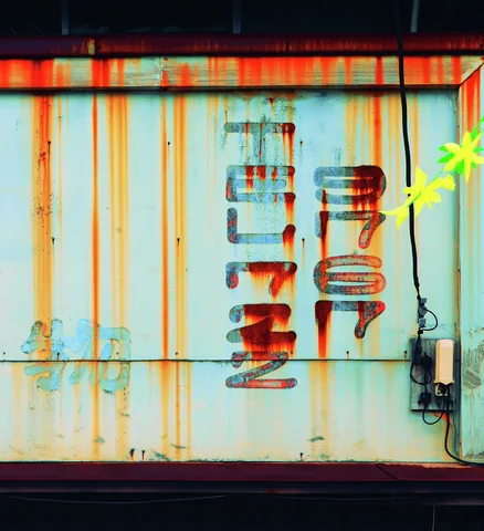 Faded Japanese characters and a phone number sit beneath orange rust streaks on a pale metal sign.