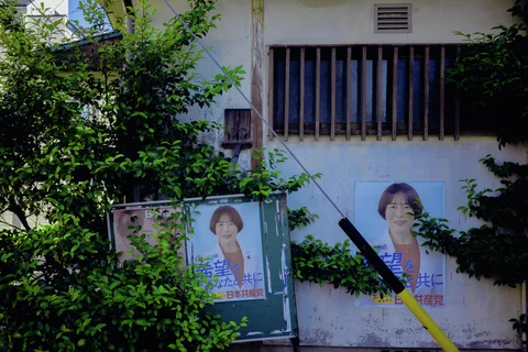 Green foliage partly covers portrait posters beneath a barred window on a pale weathered wall.