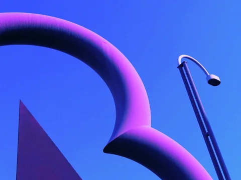 A large purple curved sculpture rises beside a slender streetlight against a deep blue sky.