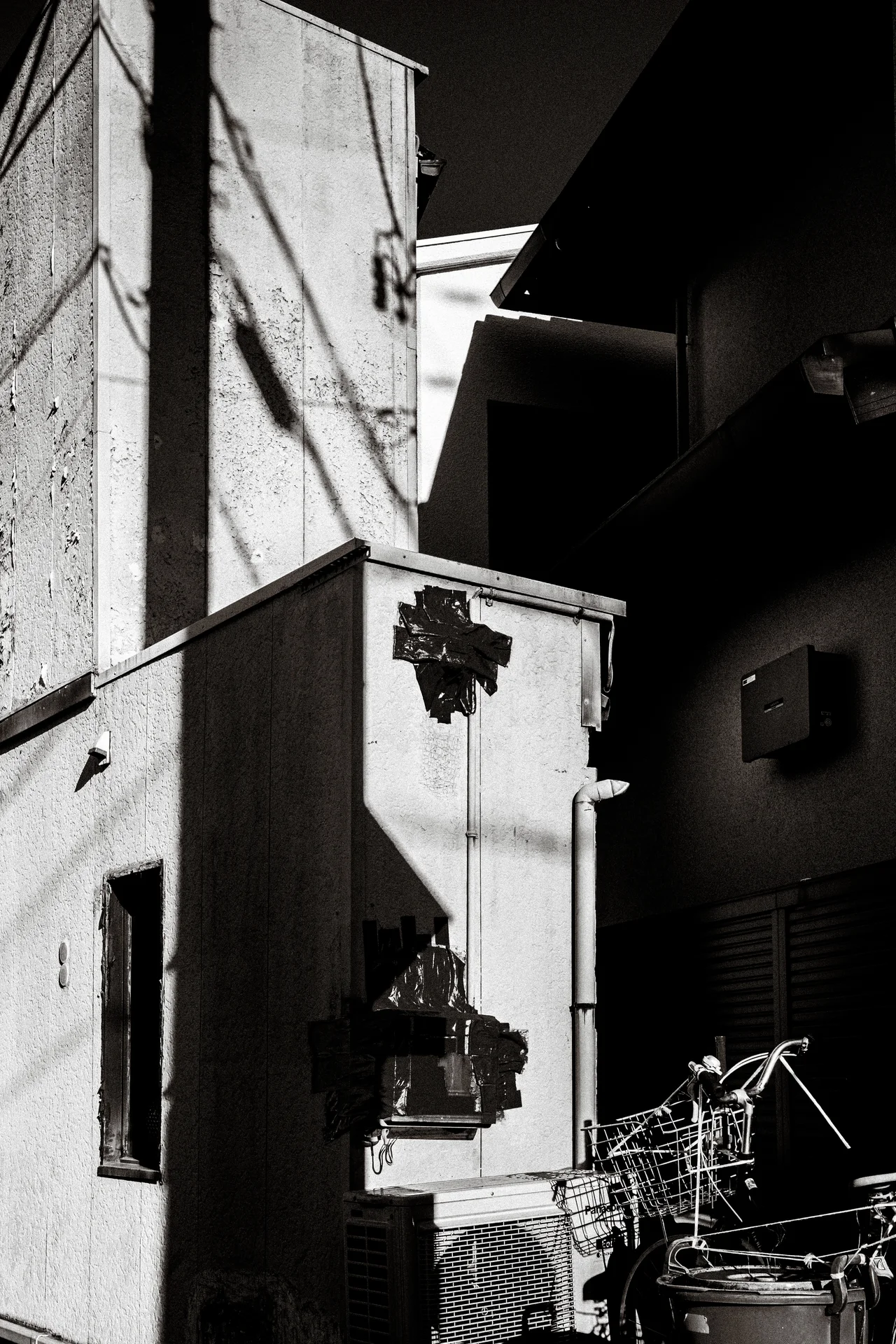 Strong shadows cross a damaged pale wall beside bicycles in a narrow monochrome passage.