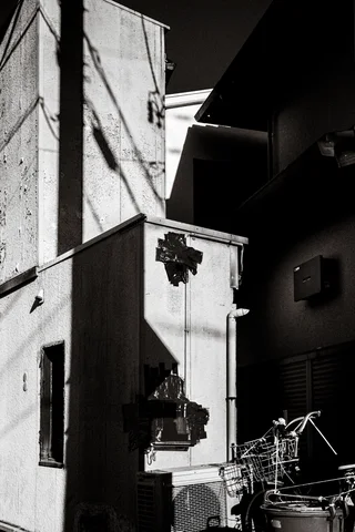 Strong shadows cross a damaged pale wall beside bicycles in a narrow monochrome passage.