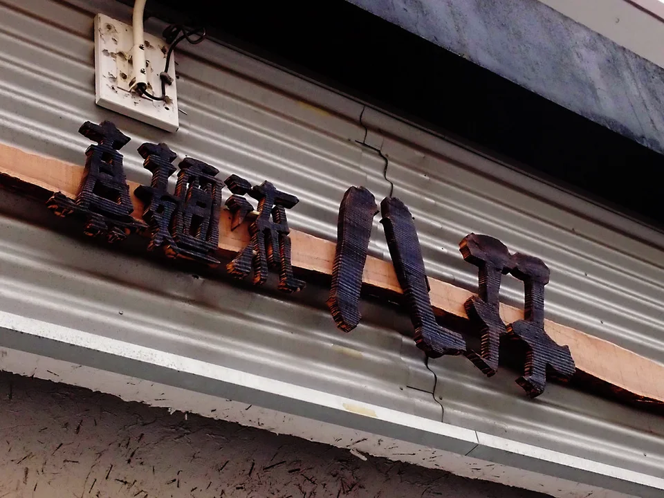 Dark raised Japanese characters attach to a rusty horizontal strip across pale corrugated siding.