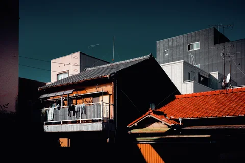 Sunlight highlights an orange balcony and red tiled roof among deeply shadowed buildings beneath dark blue sky.