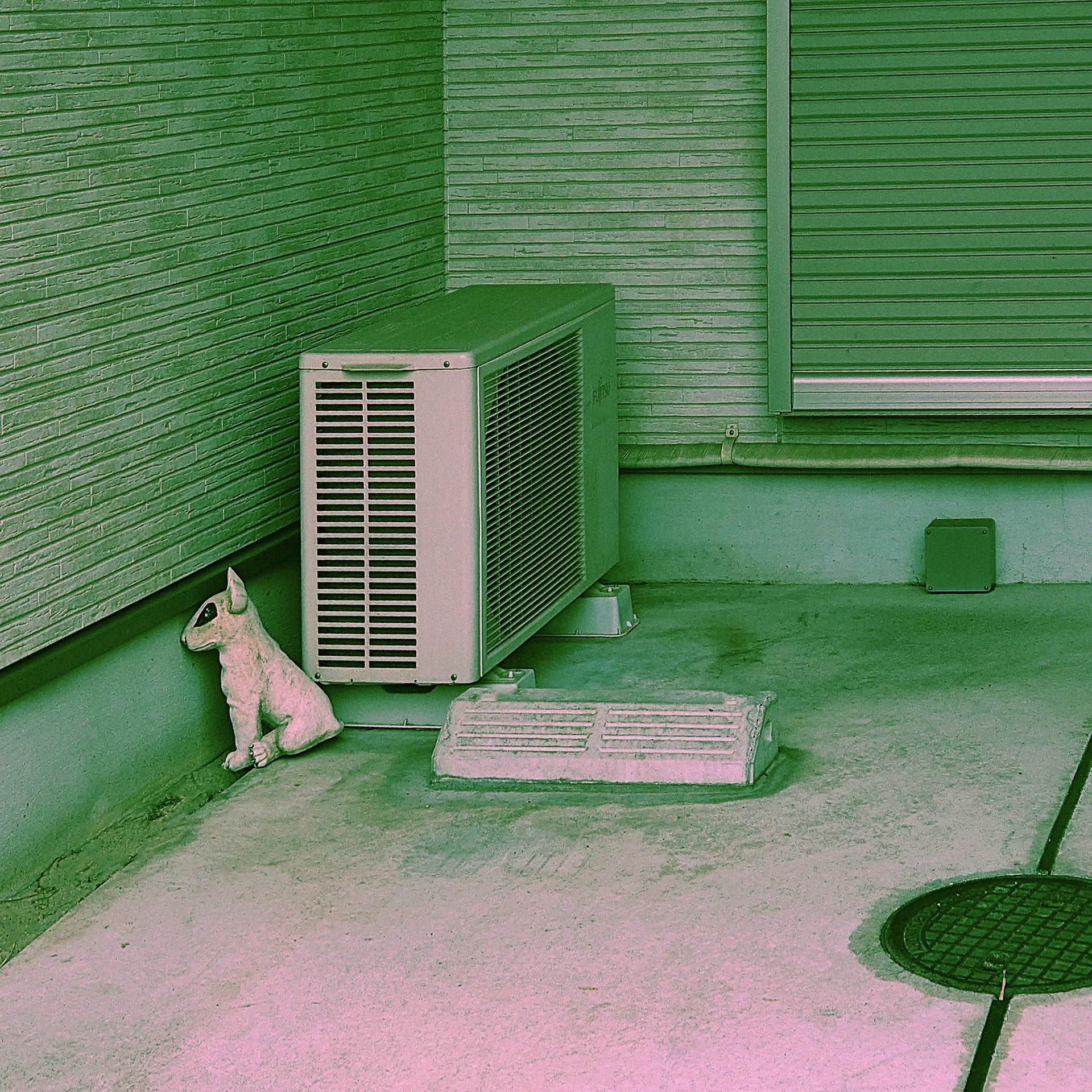 A pale cat ornament sits beside an outdoor air conditioner in a green-toned corner.