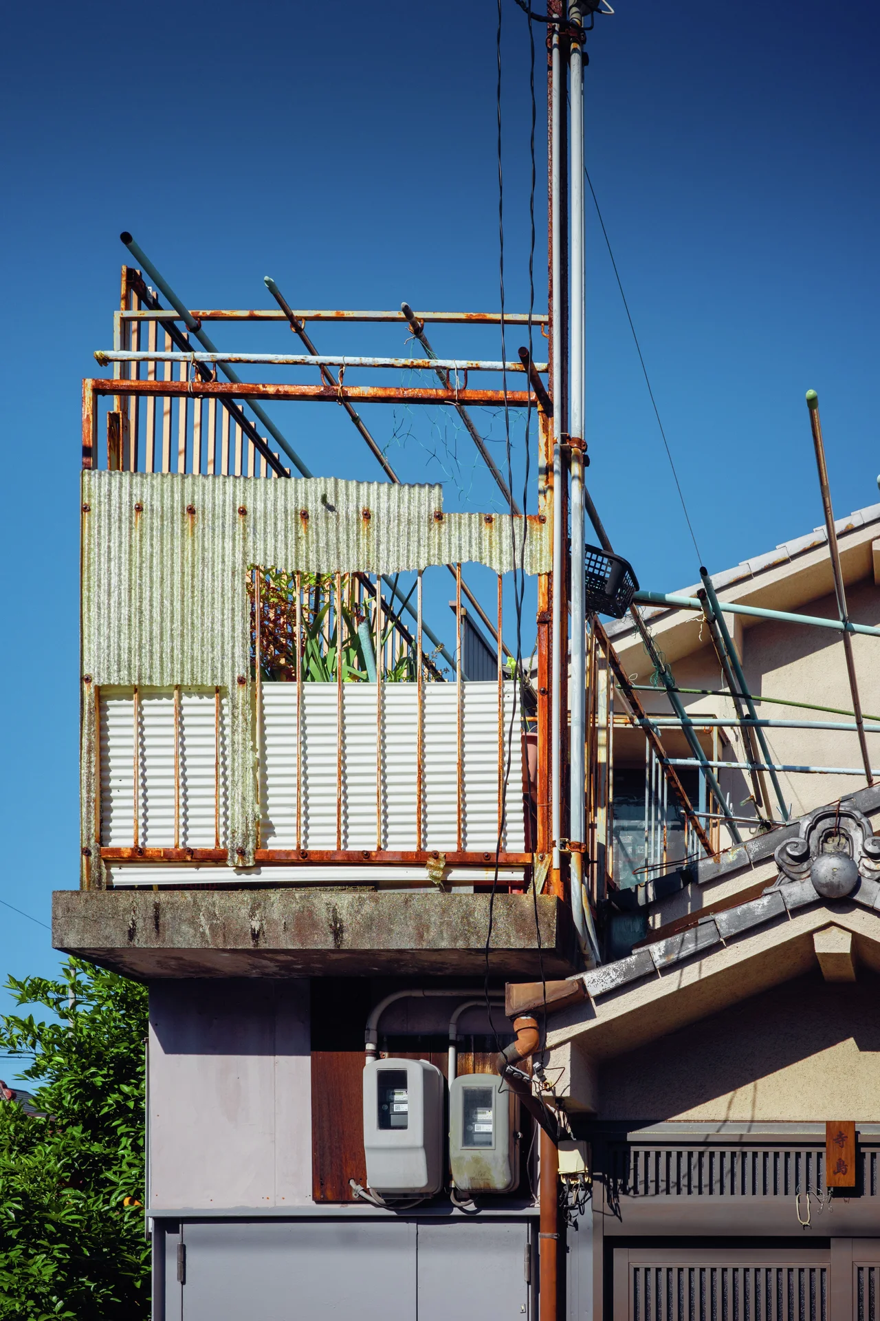 A rusty rooftop railing and bare frame rise beside utility pipes above tiled roofs and blue sky.