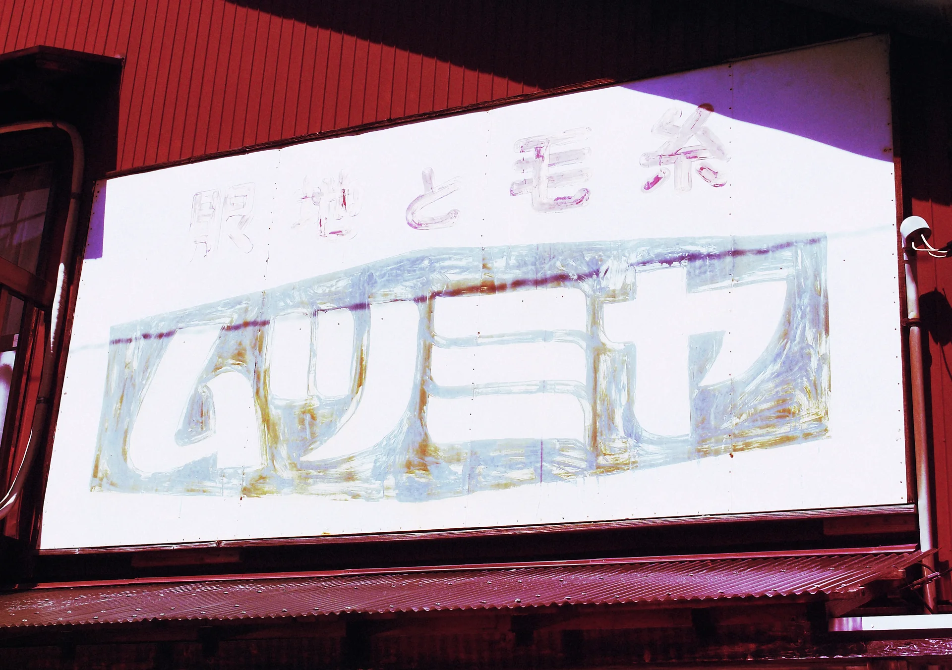A faded Japanese sign hangs above a dark red storefront awning crossed by strong diagonal sunlight.