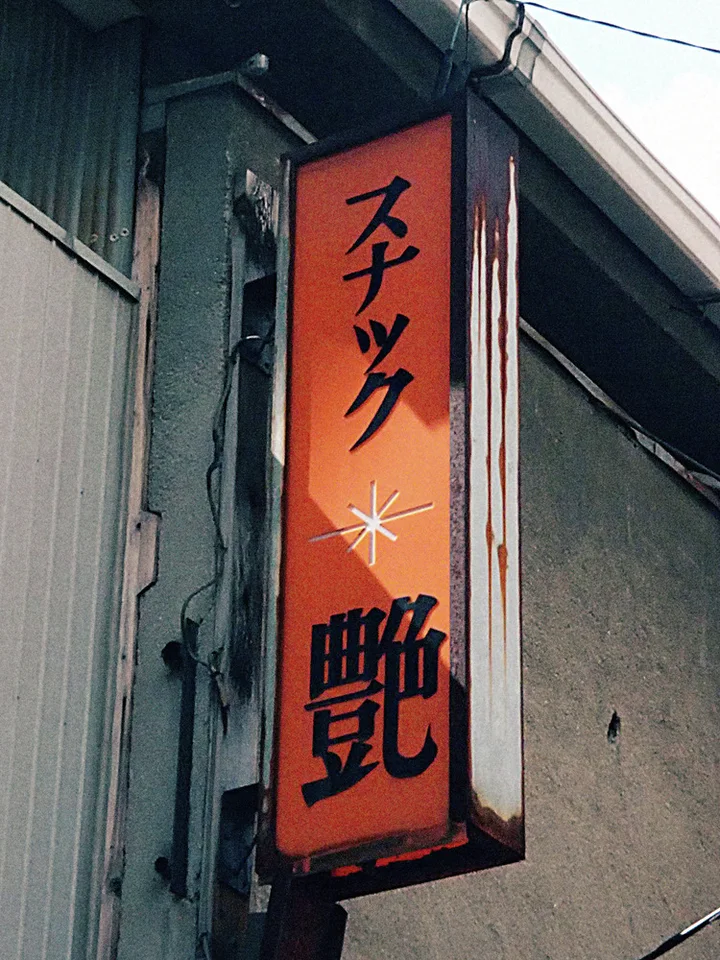 Black Japanese lettering runs down an orange projecting sign on a weathered wall beneath a roof edge.