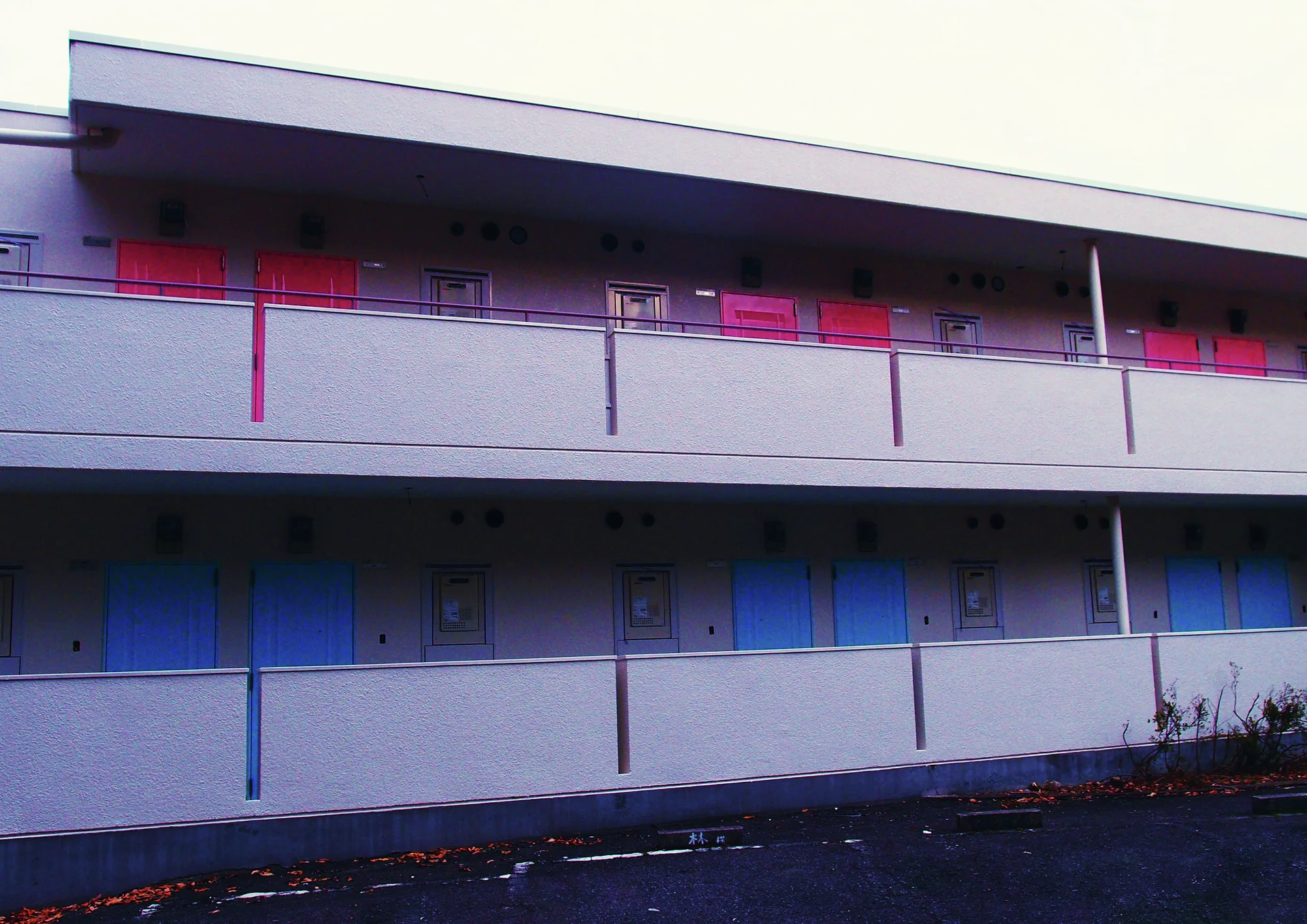 Red and blue doors repeat along two pale exterior apartment corridors bordered by simple metal railings.