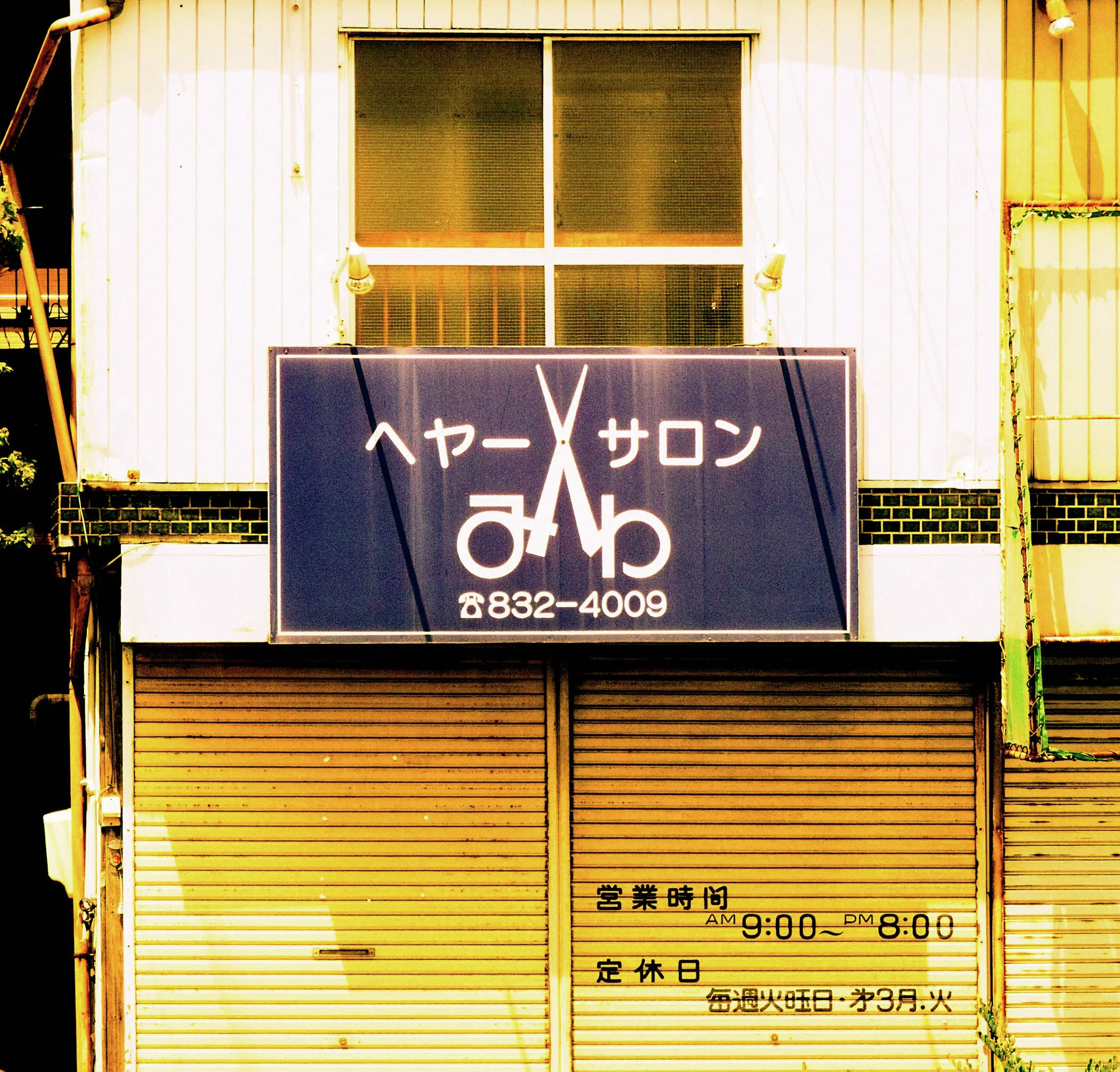 A dark salon sign with a scissors graphic hangs above closed yellow shutters beneath a curtained window.