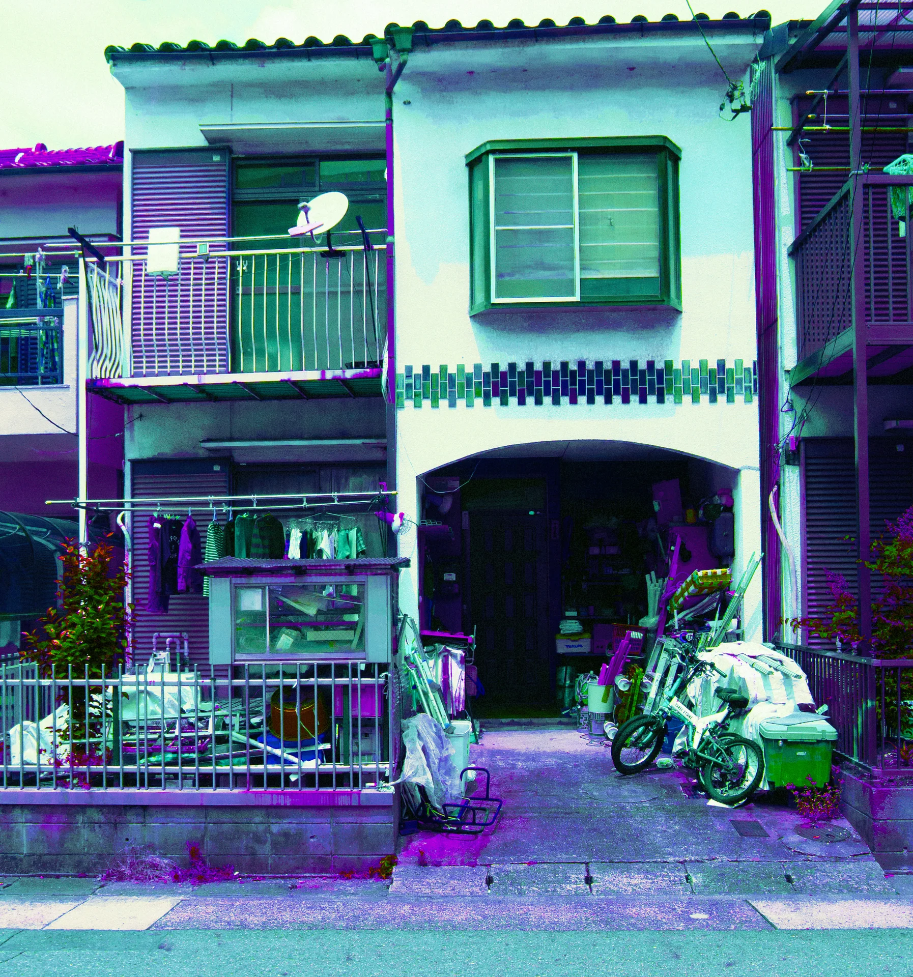 Bicycles and potted plants crowd an arched entrance beneath a narrow house balcony and curtained upper window.