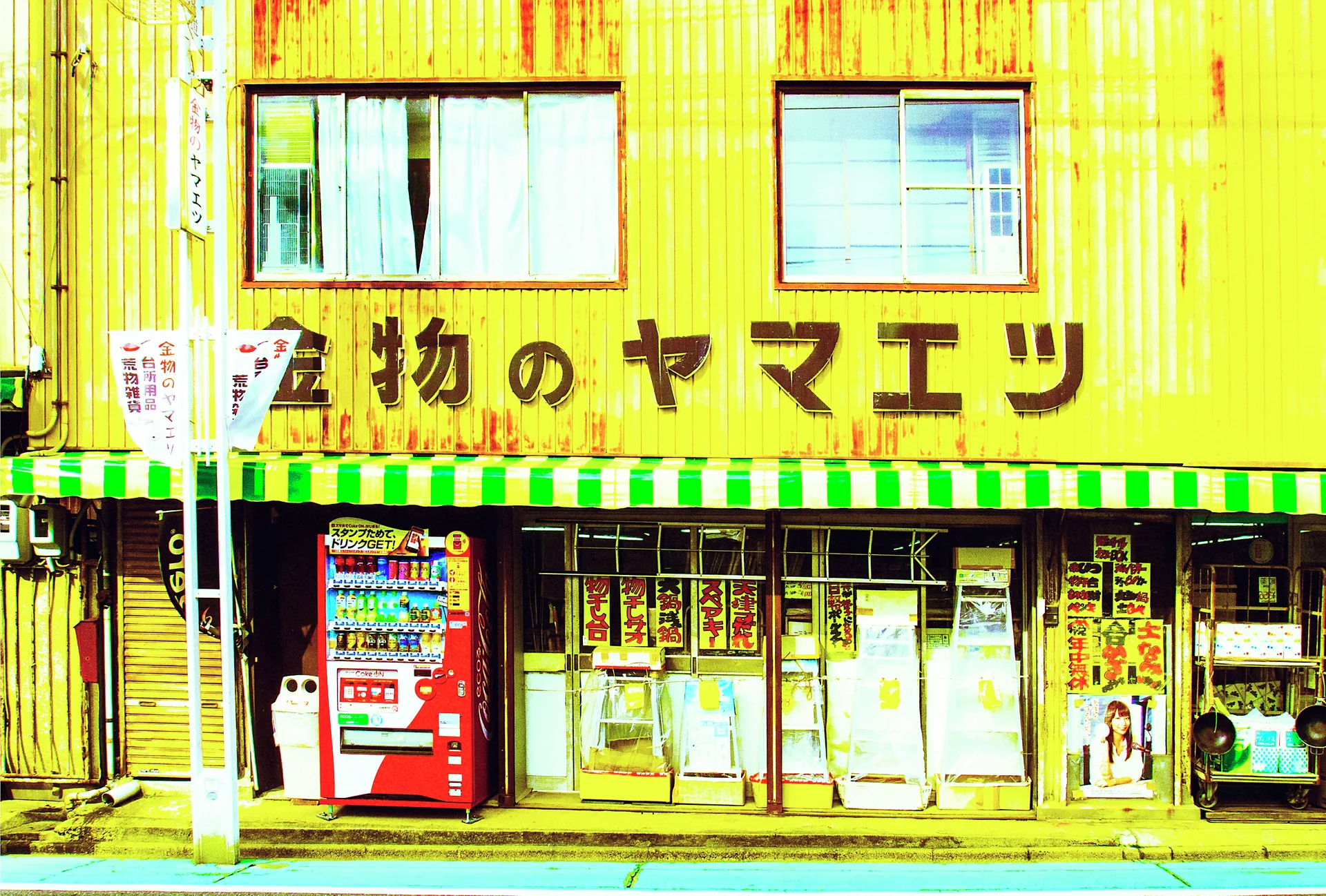 A yellow shop with green-striped awnings displays Japanese signage above glass doors and a red vending machine.
