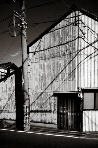 Utility wires cast angular shadows across a corrugated building beside a concrete pole in black and white.