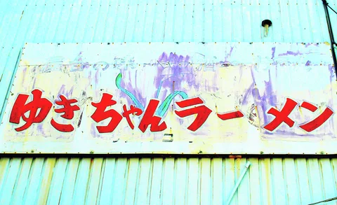 Bold red Japanese lettering crosses a peeling pale sign on turquoise corrugated siding.