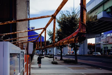 Rusty overhead pipes extend above a sidewalk where a person walks beside an elevated roadway.