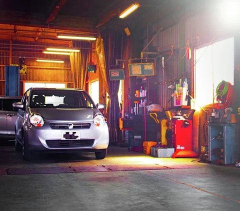 A silver car faces outward inside a workshop lit by fluorescent fixtures and warm sunlight.