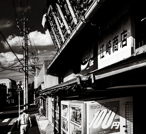Shop signs and vending machines line a shadowy street beneath dense overhead wires in black and white.