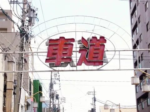 Two large red Japanese characters hang inside an arched metal gateway above a street crowded with wires.
