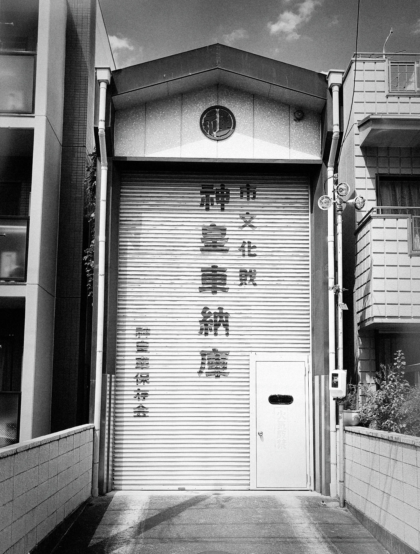 Vertical Japanese lettering covers a tall closed shutter beneath a small round window in black and white.