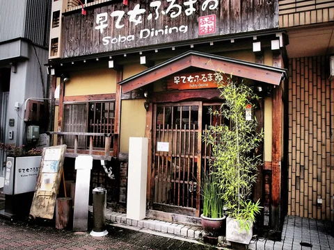 Bamboo plants flank a wooden restaurant entrance beneath a small gabled canopy and rustic signage.