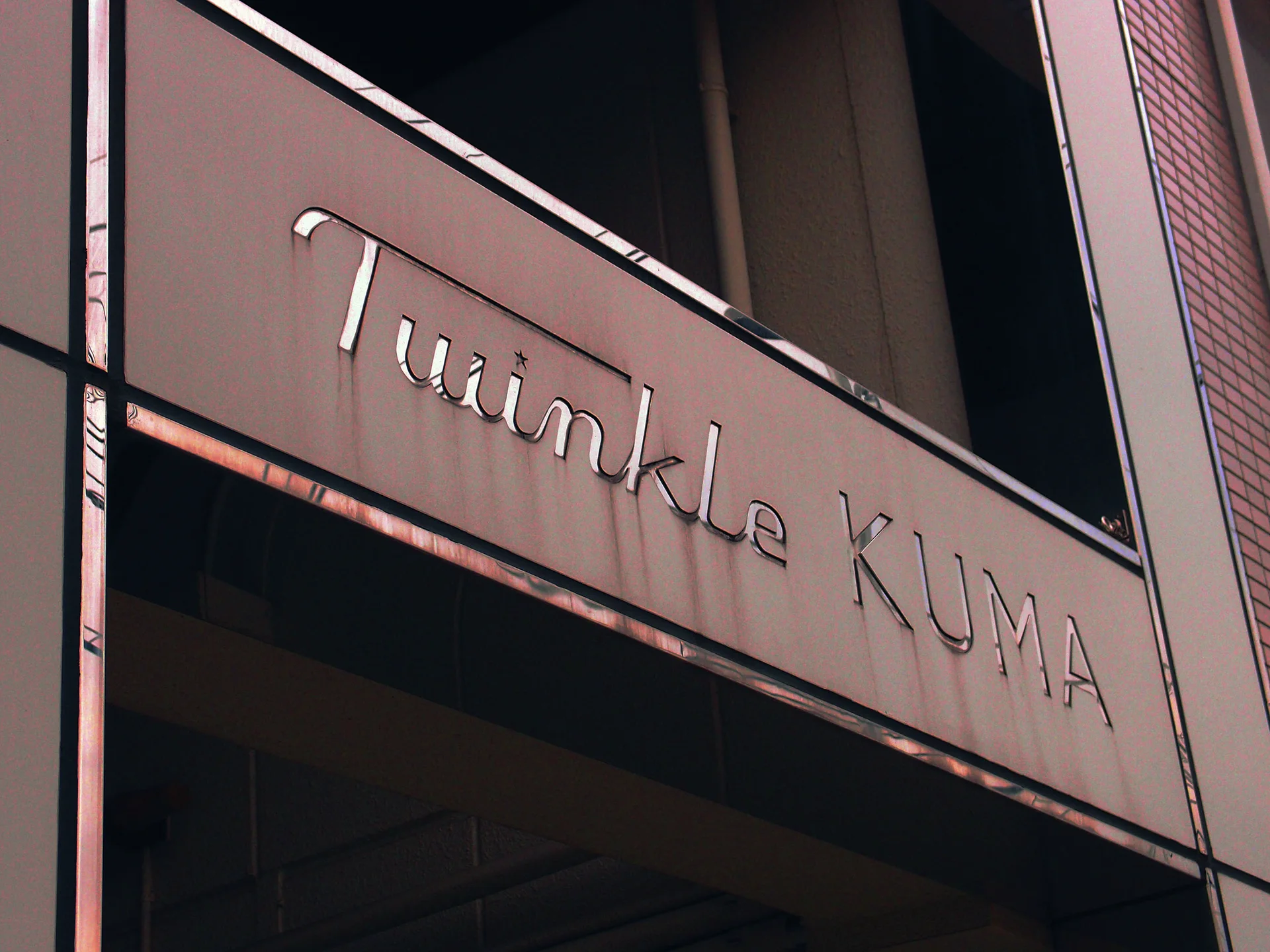Slim raised Twinkle Kuma lettering crosses a dark building panel beneath tall recessed windows.