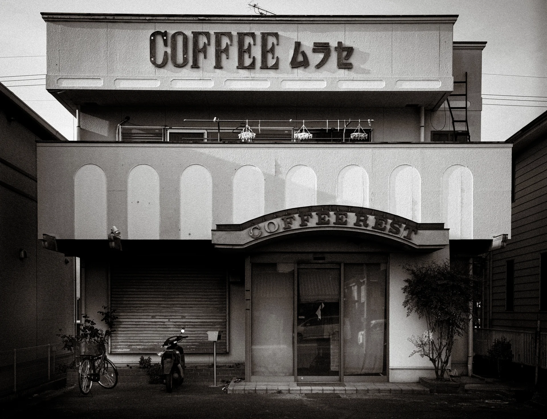 A two-story coffee shop has an arched entrance canopy and pale vertical panels above a closed shutter.