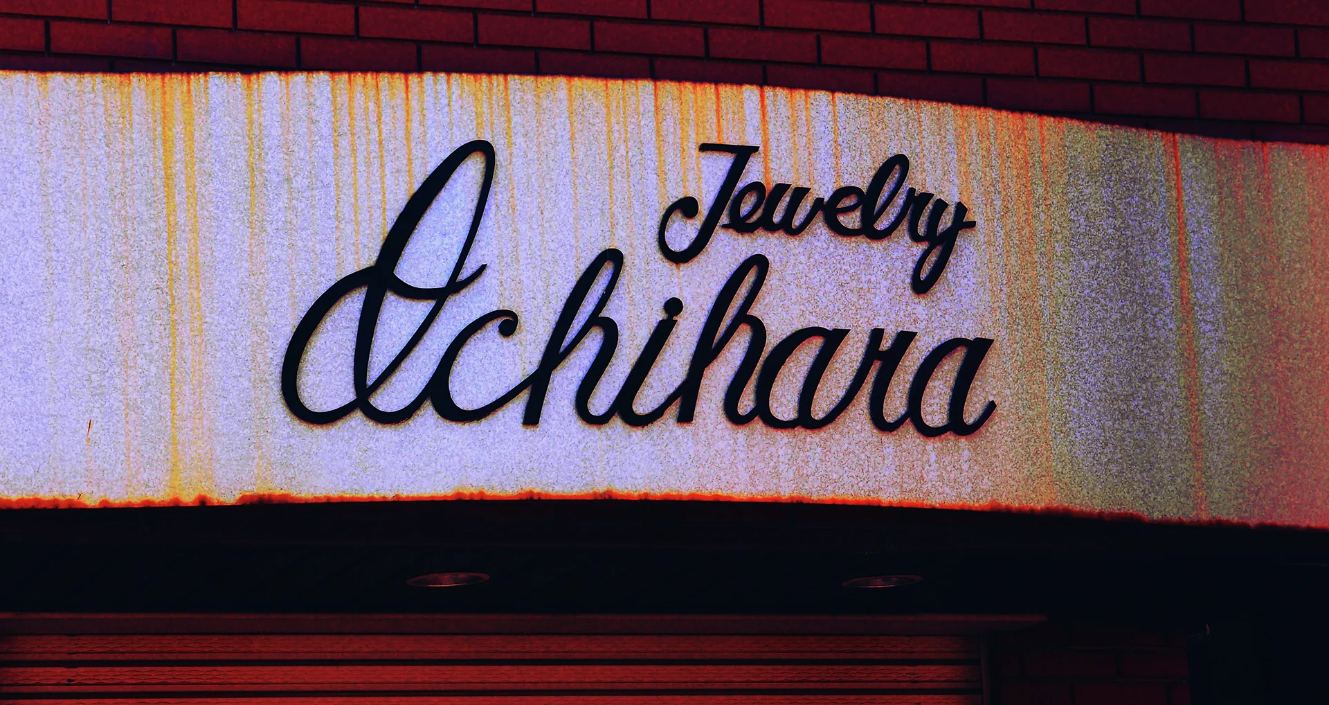 Black cursive jewelry-shop lettering spans a pale rust-streaked sign beneath a red brick wall.