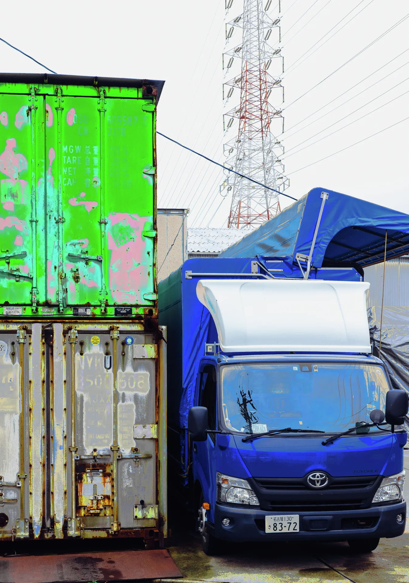 A small blue truck stands beside stacked weathered shipping containers beneath a distant electricity pylon.