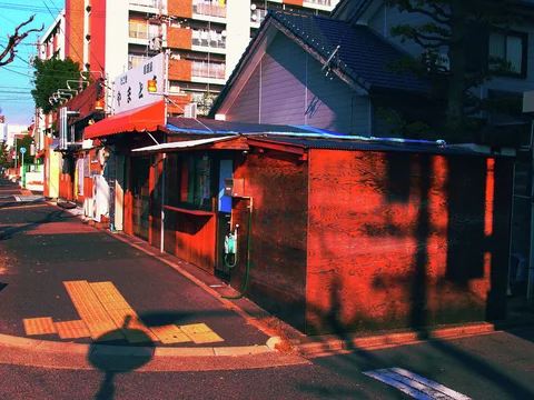 Strong shadows cross a narrow sidewalk beside weathered red-toned walls and low shop awnings.