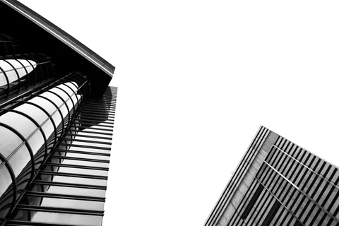 Curved glass tubes and striped building edges frame a broad pale sky in a black and white upward view.