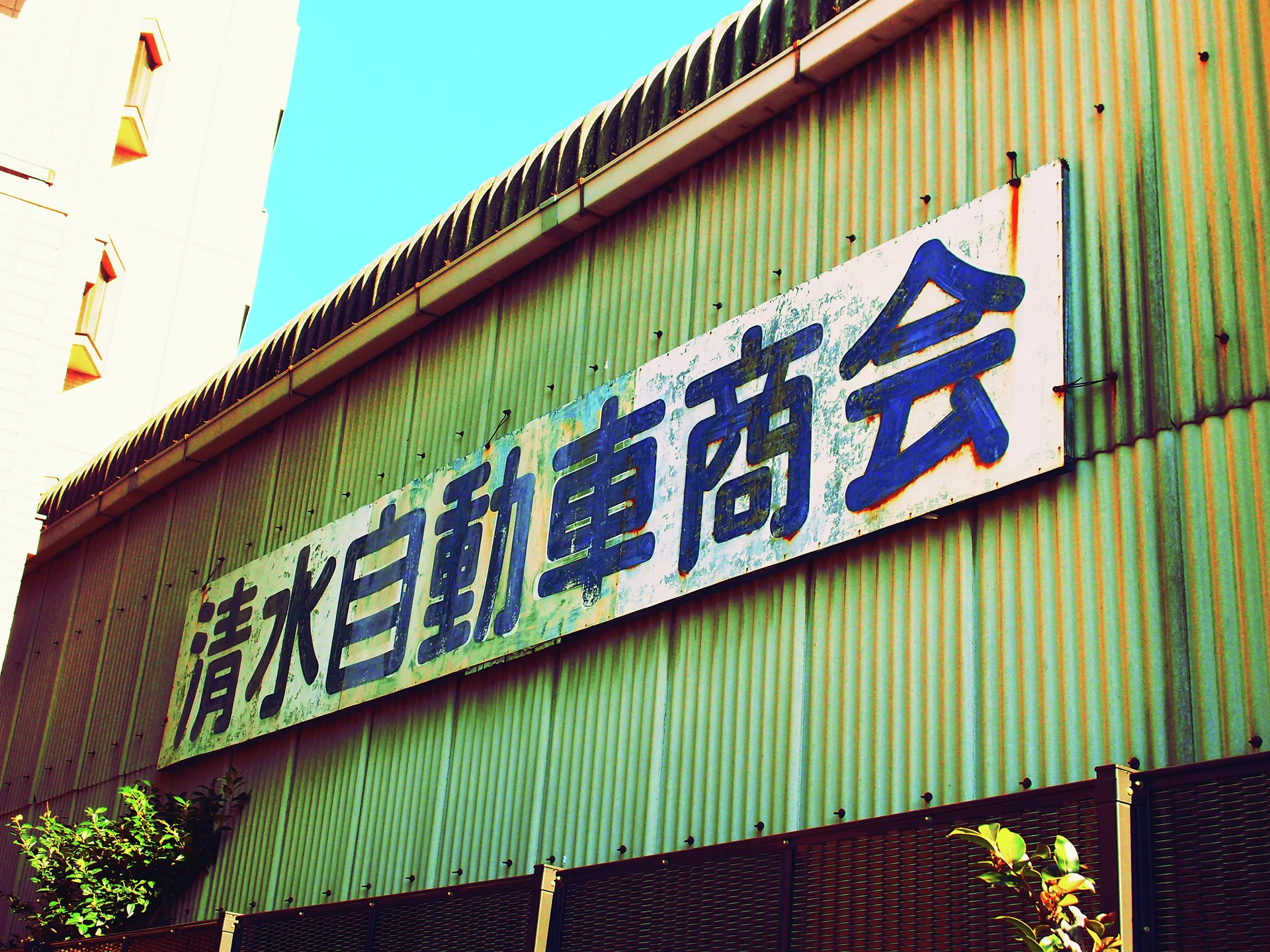 A long weathered sign with large blue characters spans green corrugated siding beneath a curved roof edge.