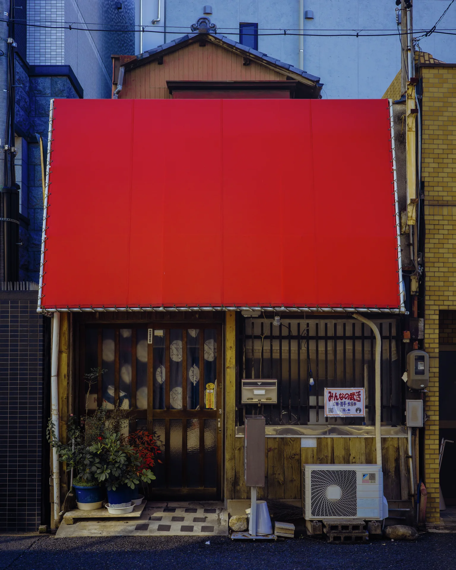 A large blank red sign dominates a narrow storefront with weathered panels and potted plants below.