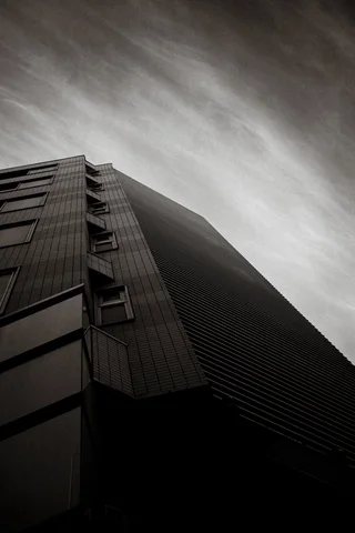 A steep upward view follows a tall louvered building toward streaked sky in black and white.