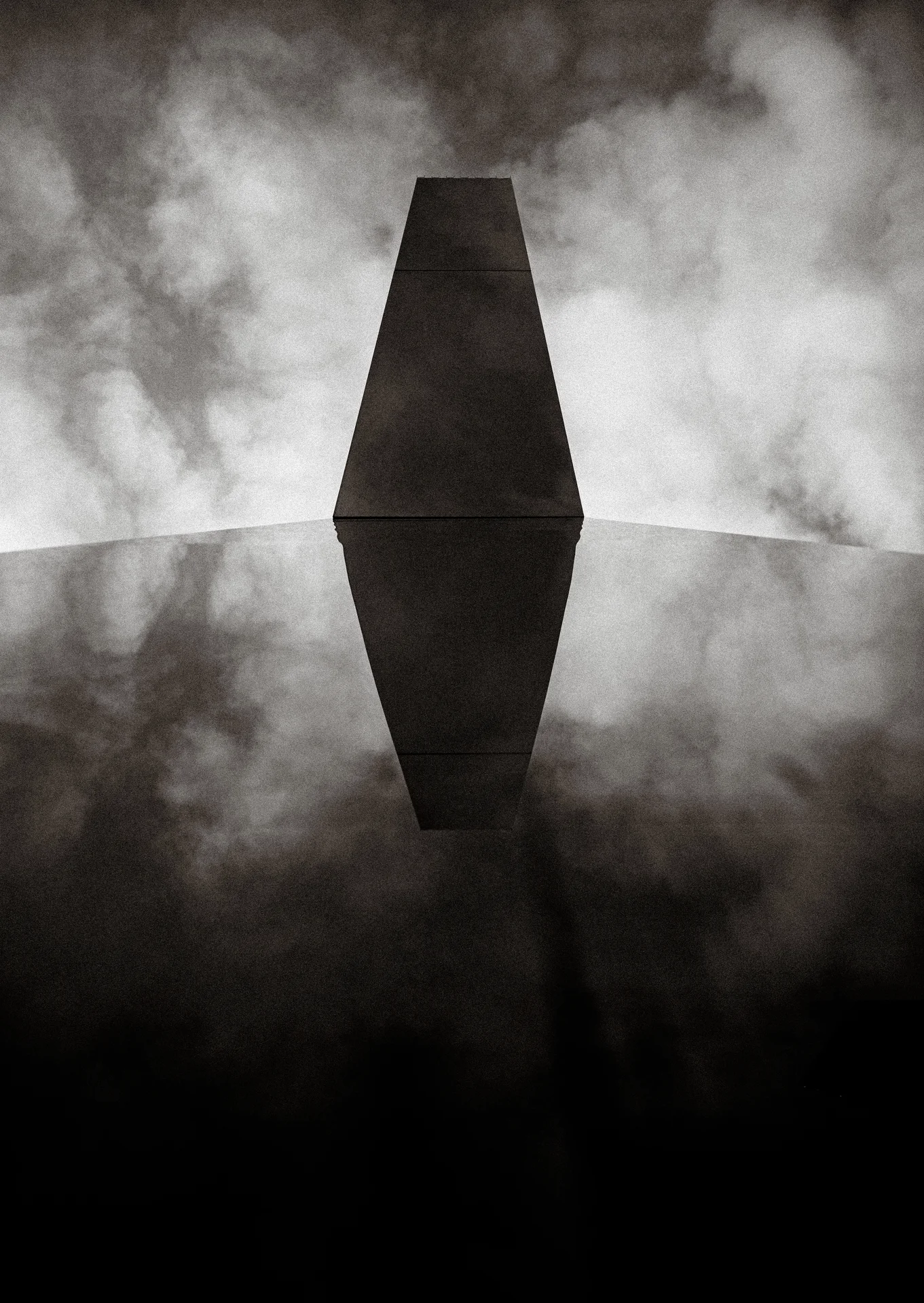 A dark tapered geometric structure meets its reflection amid cloudy light in black and white.