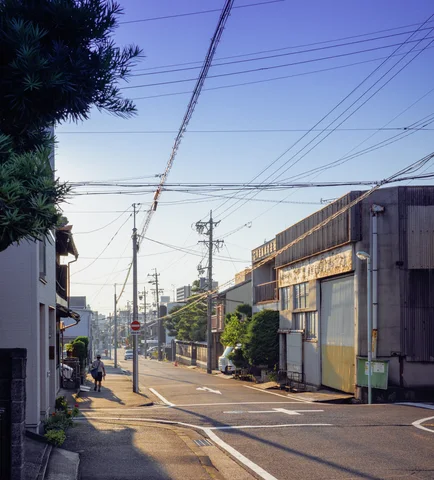 A quiet industrial street runs past low warehouses and utility poles beneath a web of overhead wires.