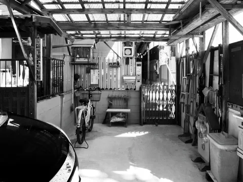 A bicycle stands inside a cluttered covered courtyard beneath translucent roof panels and exposed timber beams.