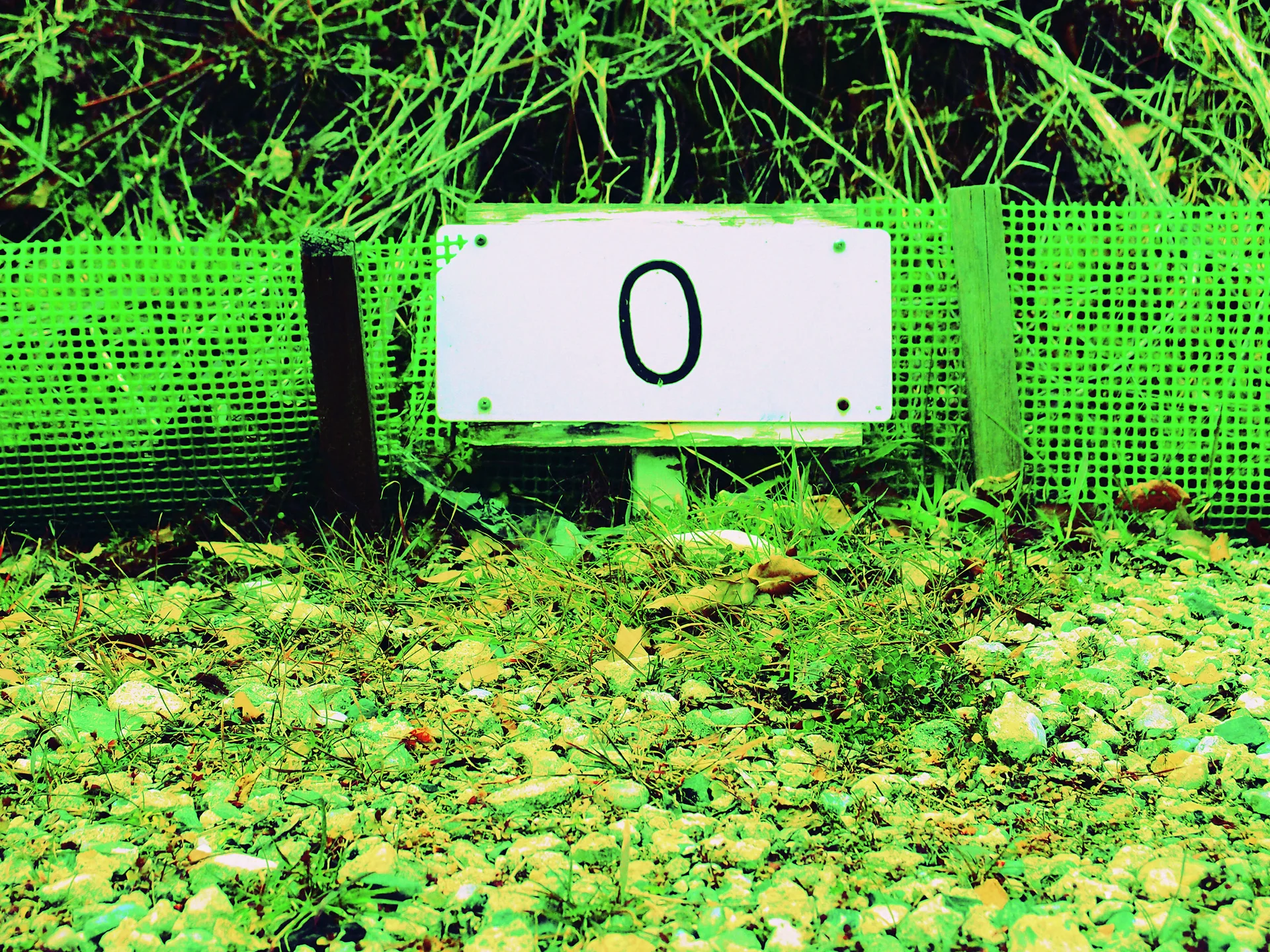 A white rectangular marker bearing a black zero stands against green mesh beside gravel and tall grass.