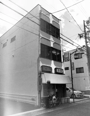 A tall narrow white building has dark windows and bicycles beside its entrance in black and white.