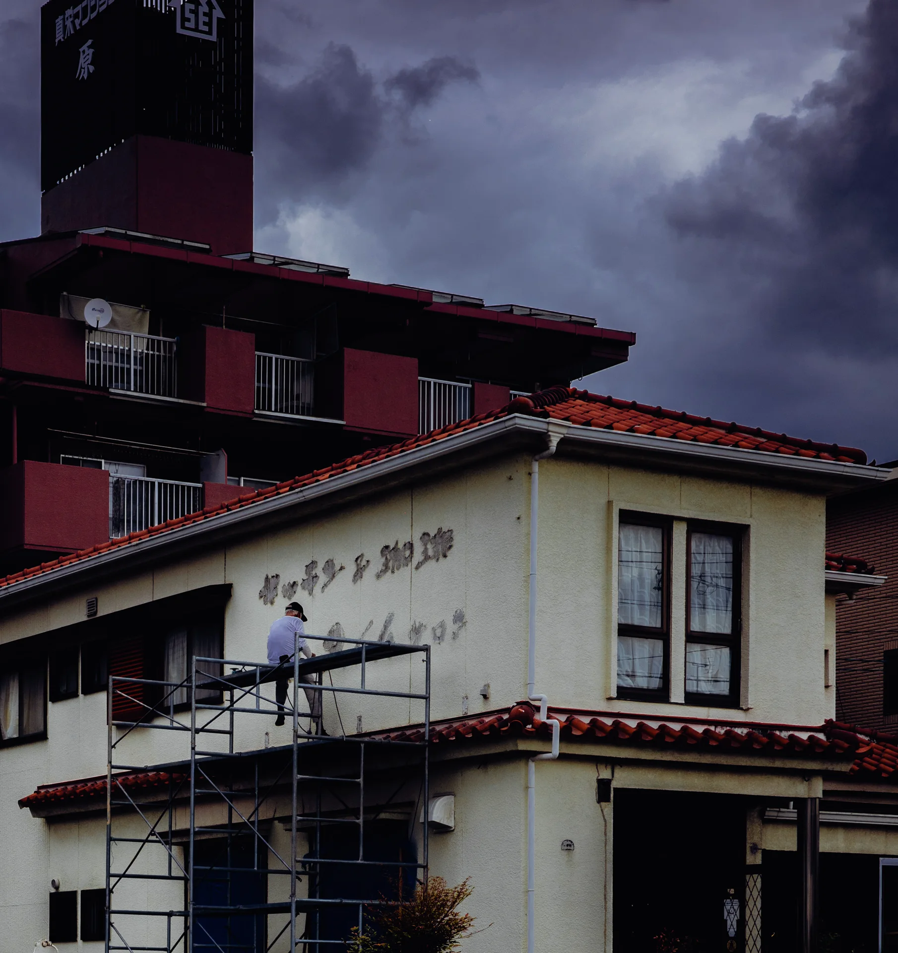 A pale building with red tiled roofs stands before a darker tower beneath heavy gray clouds.