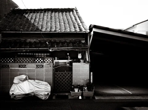 A covered scooter rests outside a tiled-roof house beside a dark open garage in black and white.