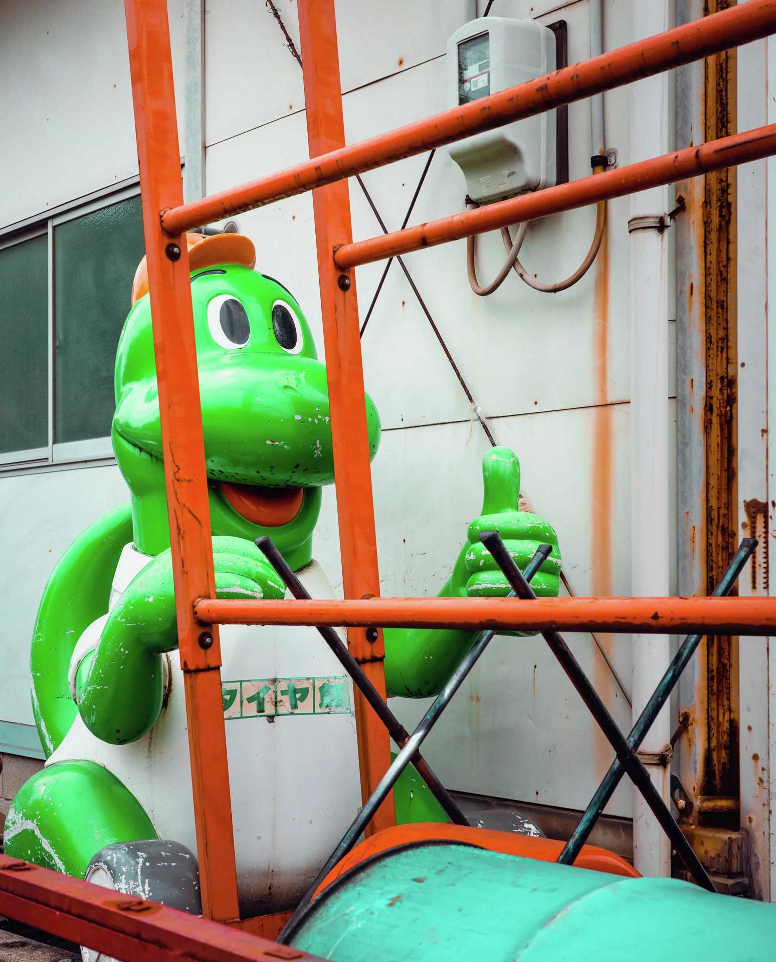 A smiling green character statue in white overalls stands behind orange metal railings.