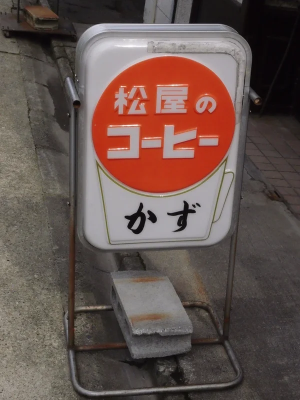 A freestanding pale coffee sign displays white Japanese lettering inside an orange circle above black script.