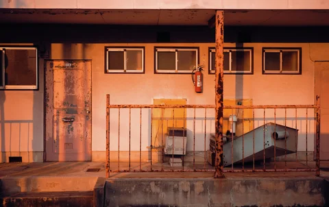 Warm light illuminates a weathered building wall with small windows behind a rusty metal railing.