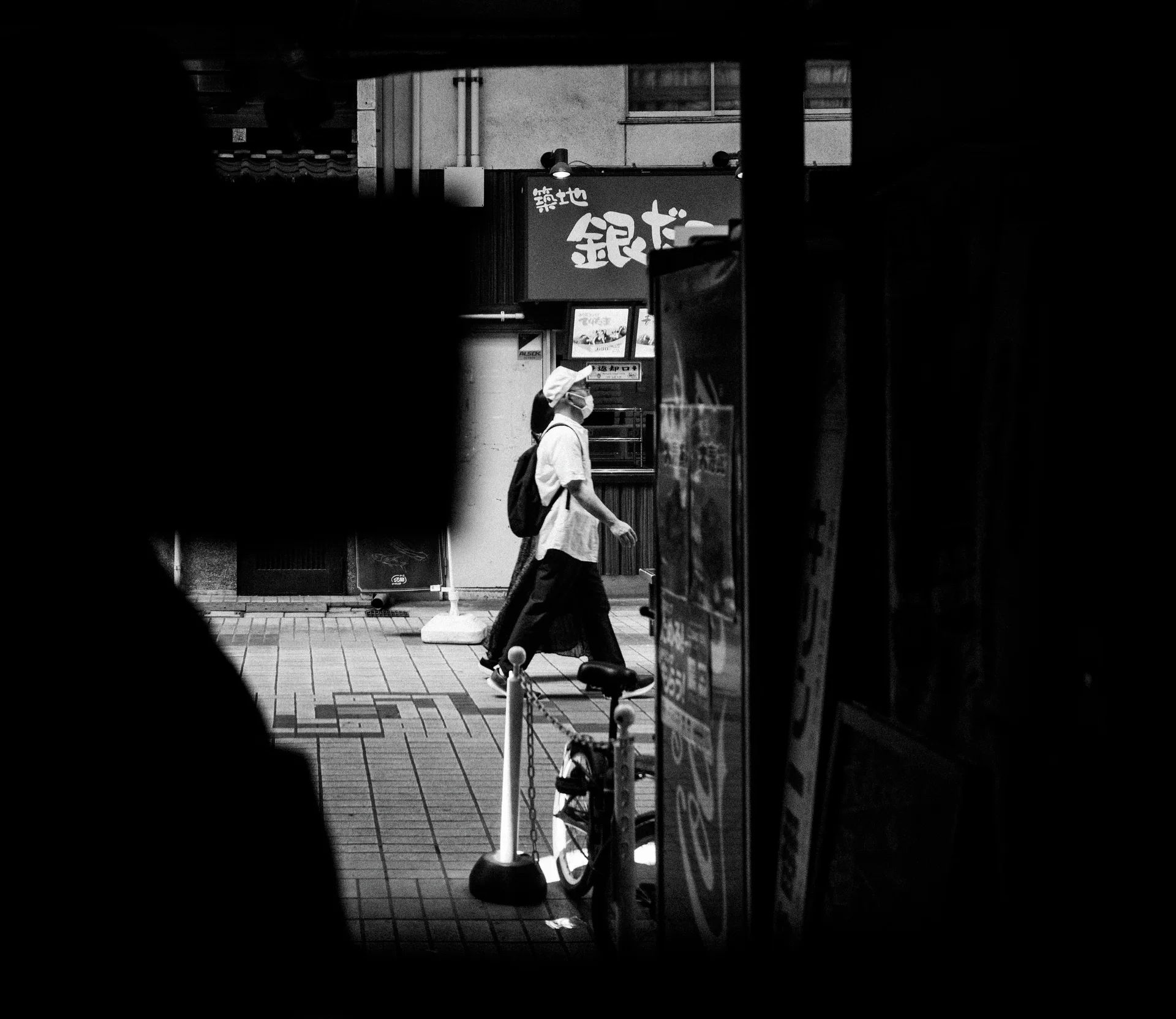 A pedestrian in light clothing passes a sunlit opening surrounded by deep shadows in black and white.