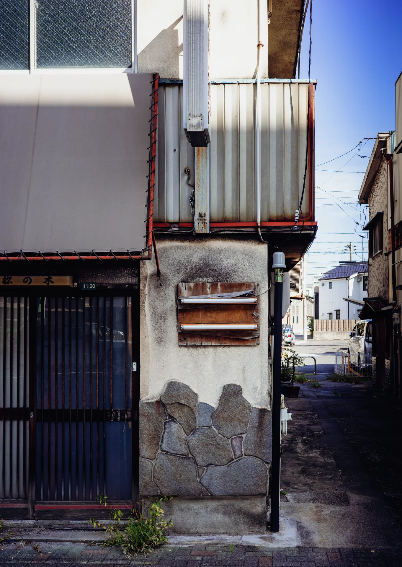 Peeling plaster surrounds a dark slatted doorway beside a narrow alley and small metal balcony.