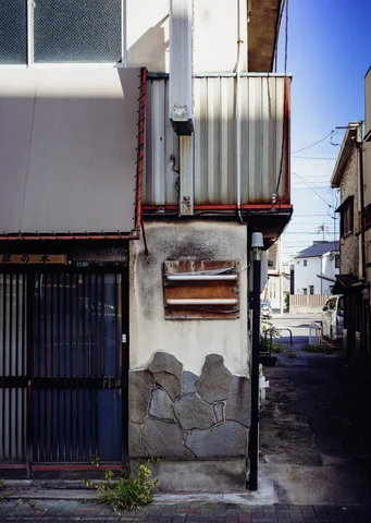 Peeling plaster surrounds a dark slatted doorway beside a narrow alley and small metal balcony.
